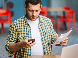 A man in yellow and blue checked shirt looks down at his phone as he holds a newspaper