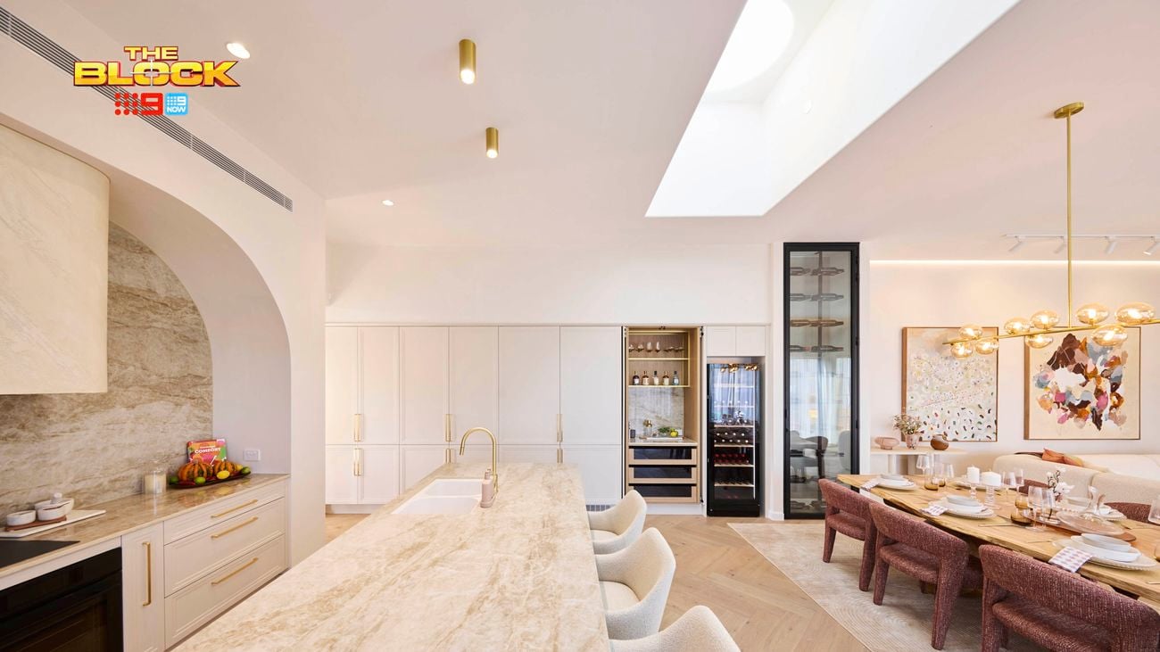 Open-plan kitchen and dining area from The Block, featuring a marble island with bar stools, built-in appliances, wine fridge, and a dining table with pink chairs under a modern gold light fixture.