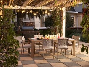   Outdoor dining area under a pergola decorated with hanging string lights, featuring a wooden table set with candles, plates, and drinks, surrounded by woven-back chairs on a tiled patio, with a barbecue grill in the background and greenery framing the space.