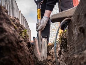 A close-up, ground-level view of a person using a metal spade to dig a trench outdoors, with soil on either side and part of a wheelbarrow visible in the foreground.