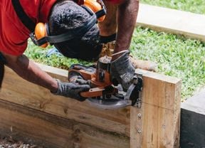 An image showing a tradesperson in PPE with a small circular saw trimming down timber sleepers for a retaining wall