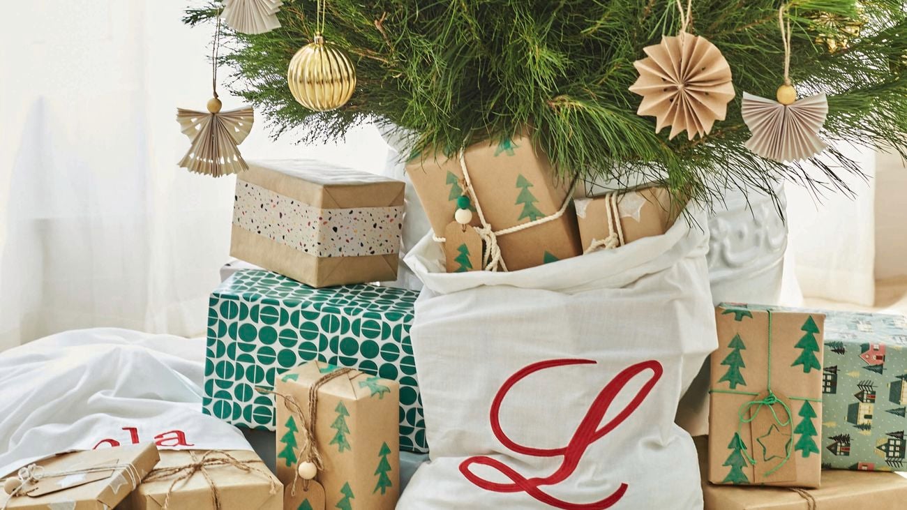 Presents wrapped in brown paper with green Christmas trees and labelled with handmade gift tags lie underneath a decorated Christmas tree.