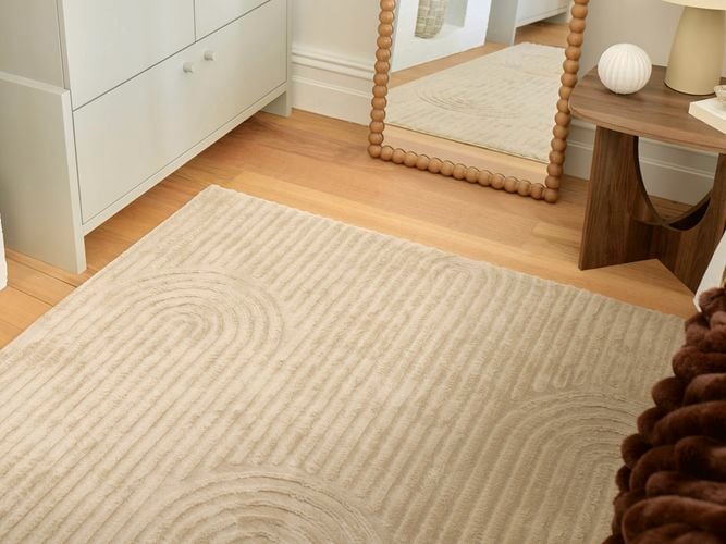 Beige textured area rug with subtle curved line patterns on a light timber floor, styled beside a cream drawer cabinet, beaded mirror, and wooden side table in a minimalist bedroom.