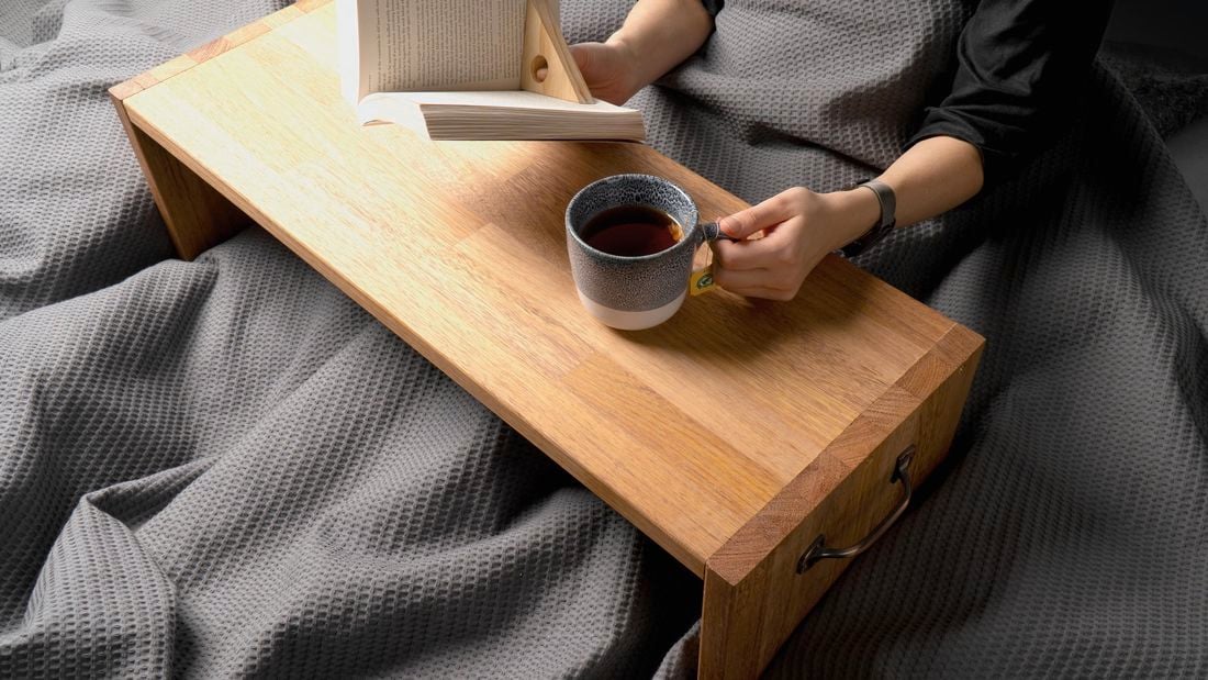 A pair of hands resting on a timber breakfast table holding a book and cup of tea from bed