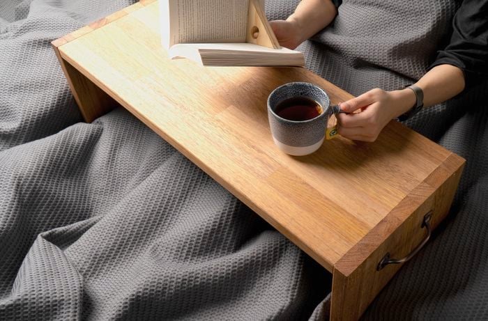 A pair of hands resting on a timber breakfast table holding a book and cup of tea from bed