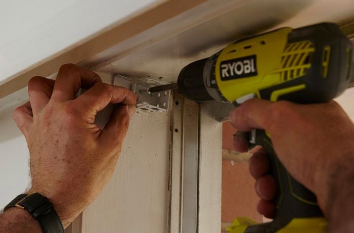 A person screwing a bracket to the side of a window cavity using a cordless drill