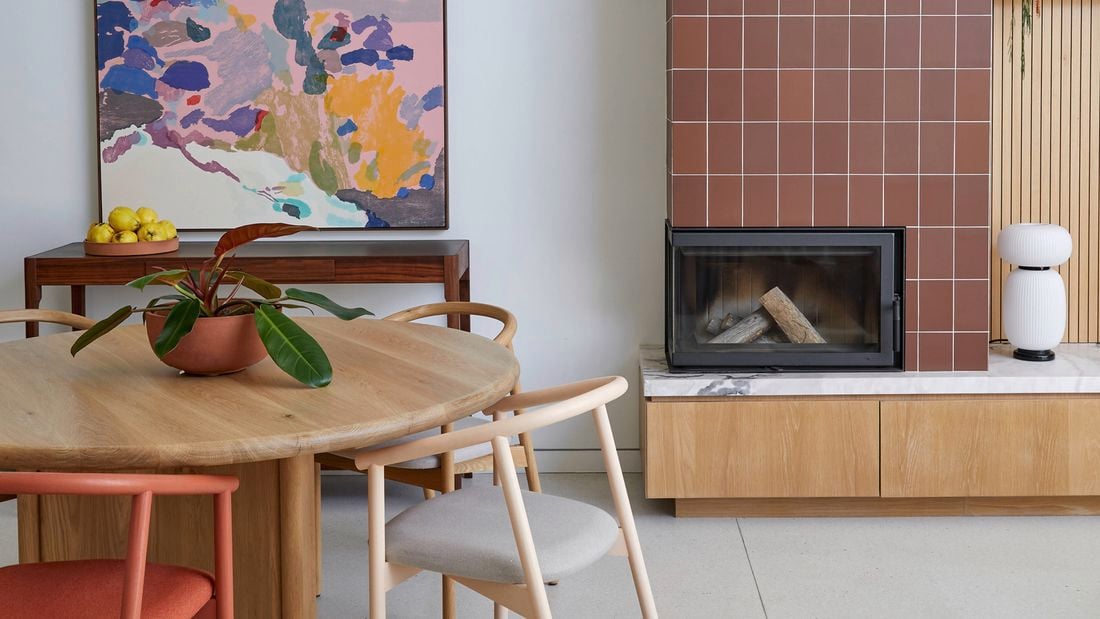 Modern dining area with a round wooden table and chairs, a white pendant light, a colourful abstract painting, and a built‑in fireplace clad in terracotta tiles with a black flue and timber base cabinet.