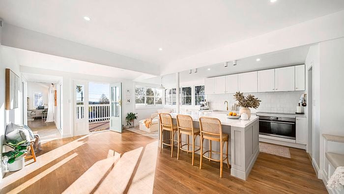 Bright open-plan kitchen and living area with a large island bench, white cabinetry, timber flooring, woven bar stools and sunlight streaming through open doors and windows.