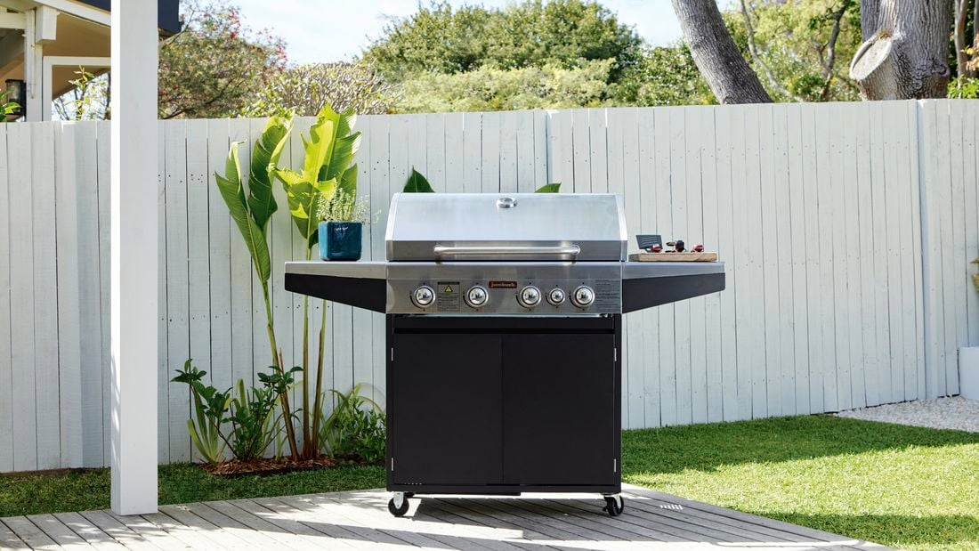 Barbecue sitting on deck with lid closed, white fence and plants are seen in the background.