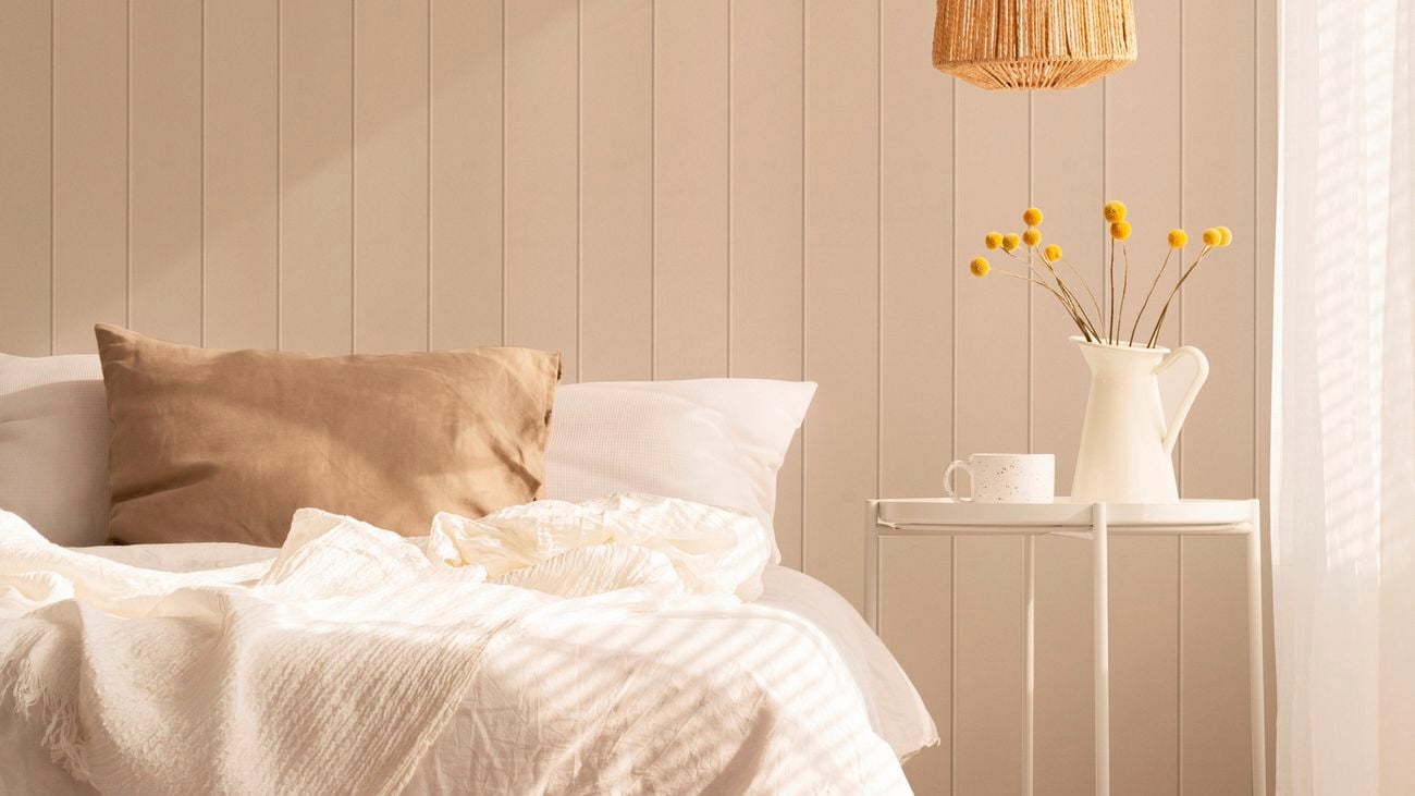 Bedroom with light beige vertical panelled walls, a bed with white bedding and a beige cushion, a white side table holding a jug of yellow flowers and a cup, a woven rattan pendant light, and a sheer curtain on the right.