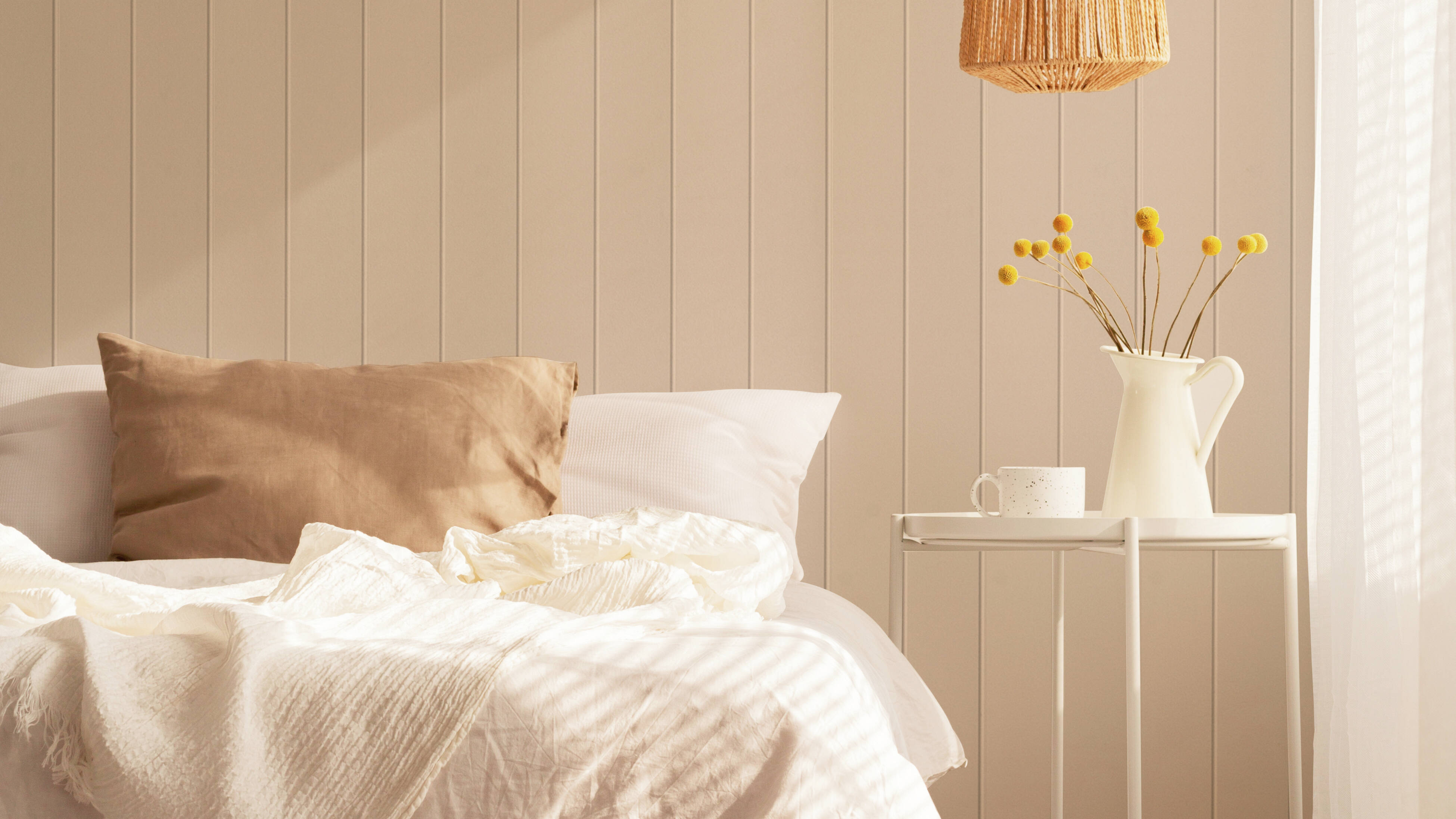 Bedroom with light beige vertical panelled walls, a bed with white bedding and a beige cushion, a white side table holding a jug of yellow flowers and a cup, a woven rattan pendant light, and a sheer curtain on the right.