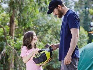 Father and daughter standing next to a tent at a campsite holding a Ryobi radio speaker with large trees in the background.
