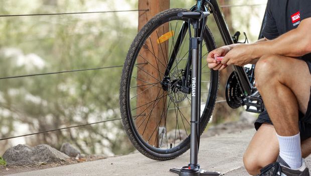 A cyclist kneels beside a road bike, adjusting its front wheel on a floor pump outdoors