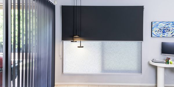 Modern interior space featuring layered window furnishings, including dark vertical blinds on a floor-to-ceiling glass door and dual roller blinds on a large window. The room has light grey walls, tiled flooring, black pendant lights, a small white desk with a computer monitor, and framed wall art, demonstrating contemporary window covering options for light and privacy control.