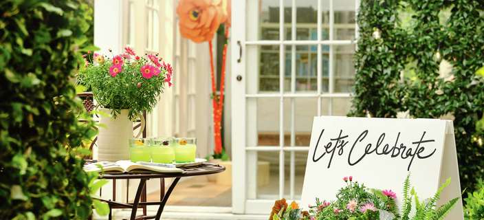 An outdoor patio with a bistro table, a planter of flowers, and a white door with a large paper flower decoration.