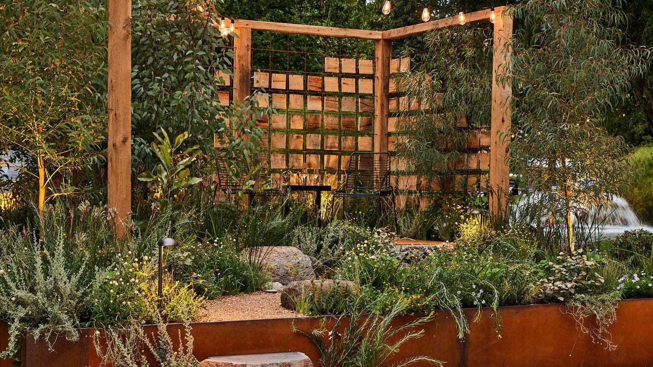 A stone step leads to a raised garden area bordered by corten steel with a timber pergola at the back. A black metal table and chairs sit on a platform with greenery all around.
