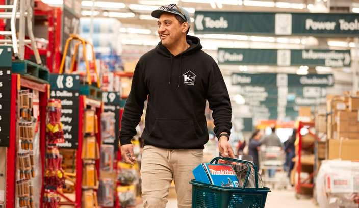 A person wearing a black hoodie and beige pants is walking through a Bunnings store carrying a green shopping basket filled with tools and products.