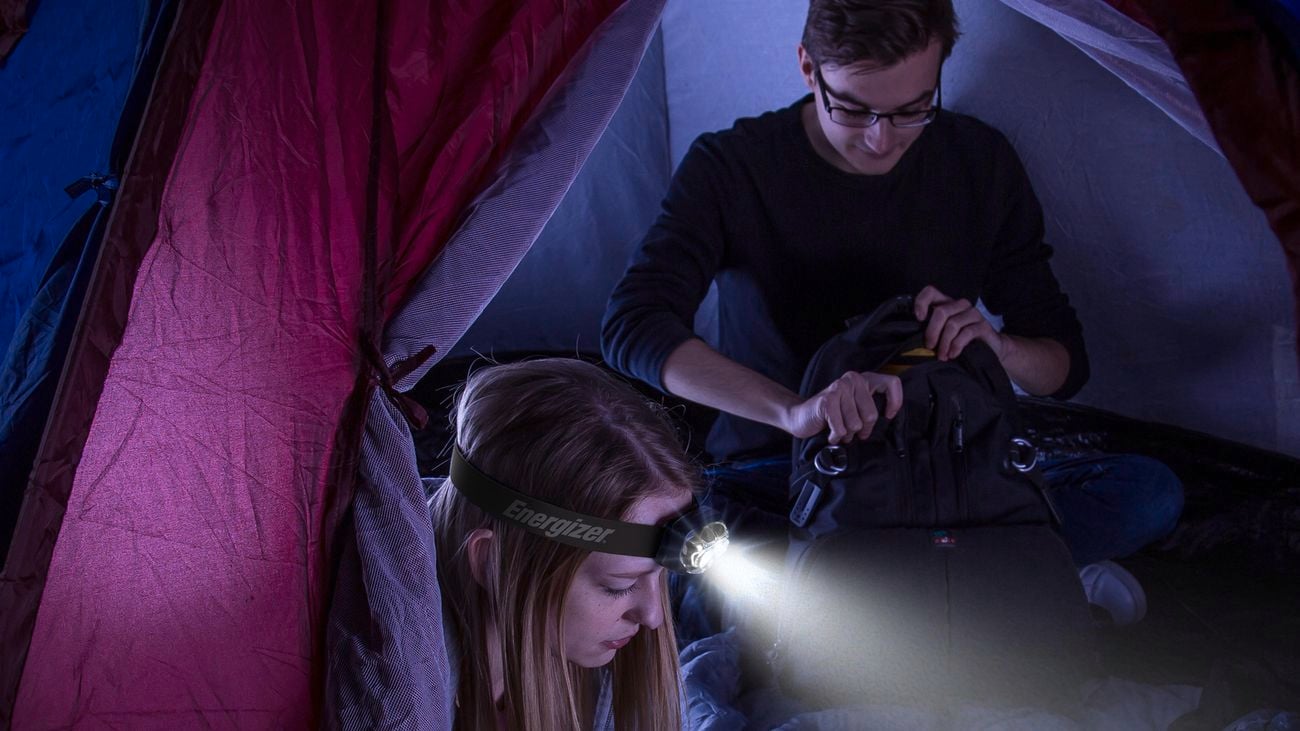  A male and a female sitting inside a tent at night. The woman is using a head torch to shine a light on the book in her hand and the man is looking through his backpack
