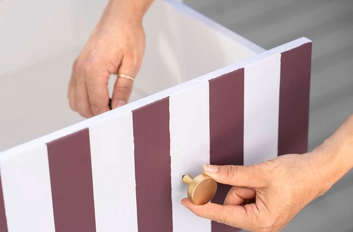 Hands fitting a round gold knob onto the front of a striped cabinet drawer.