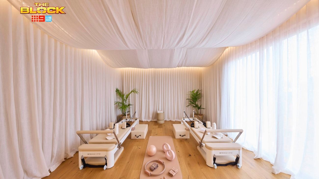An elegantly designed Pilates studio with four reformer machines arranged symmetrically. The space features white draped curtains on the walls and ceiling, creating a serene ambiance. A pink exercise mat in the center holds pink fitness accessories including an exercise ball, resistance bands, and weights. Two potted plants sit at the far end, and the floor is light-colored wood. 