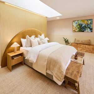 A tastefully decorated bedroom with a large bed centered under a skylight, dressed in a beige blanket and multiple pillows. Two bedside tables with lamps flank the bed. A fireplace is visible on the right, with a bench at the foot of the bed. A colorful painting hangs above a small plant and a piece of furniture. The room features neutral-toned walls and carpeted flooring.