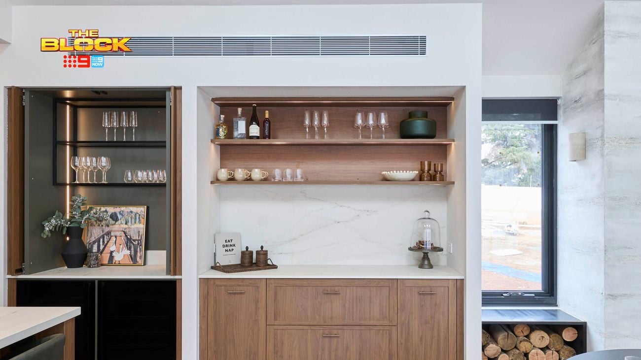 Modern kitchen bar area from The Block, featuring wooden cabinetry, open shelves with glassware and decor, a built-in wine fridge, glass cabinet, and a firewood storage nook beside a large window.