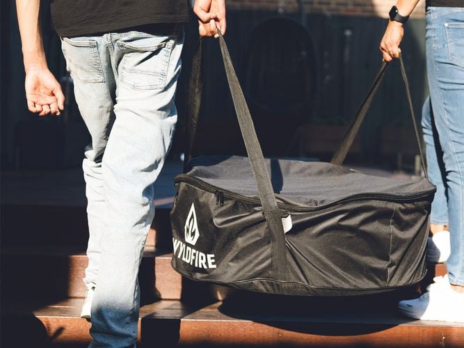 Two people carrying a black Wildfire branded carry bag containing a portable fire pit across an outdoor deck.
