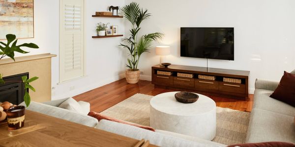 Light-filled living room with wooden floors, a wall-mounted TV above a low timber cabinet with baskets, a round coffee table on a rug, neutral sofas, indoor plants, and minimalist wall shelves.
