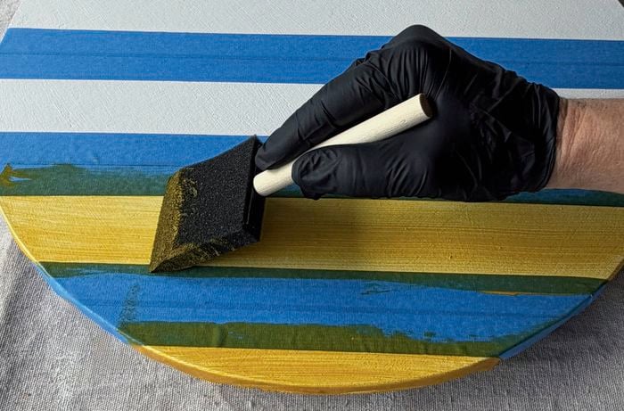 A hand in a black glove painting yellow stripes on a white-painted plywood circle, masked off with blue painter’s tape