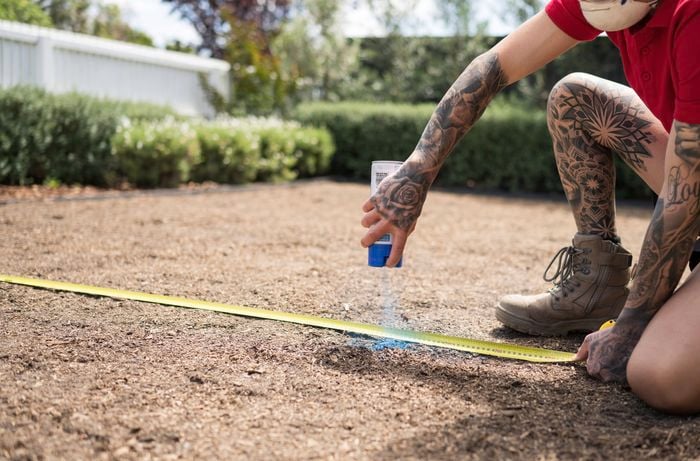 A person wearing a red shirt using a tape measure to mark the area with blue spray paint. ]    