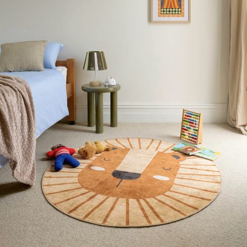 A child’s bedroom with a low bed, soft toys, a round lion-shaped rug, a small side table with a lamp, books on the floor, and daylight coming through sheer curtains.