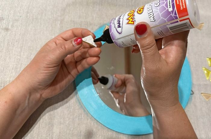 Hands applying craft glue to the back of a small triangular embellishment above a teal-painted round mirror, with more pastel embellishments scattered nearby.