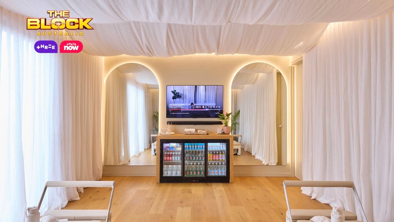 A modern Pilates studio with two reformer machines flanking a central yoga mat. The space features light wooden flooring, white curtains, and a ceiling draped with white fabric for a calming ambiance. At the far end, a mirrored wall with an arched design sits below a mounted TV and a cabinet stocked with beverages. 