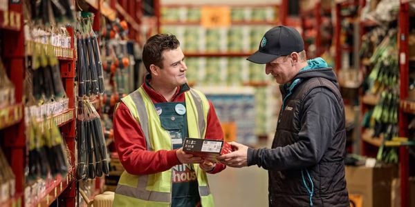 Bunnings team member handing a boxed product to a customer in a hardware store aisle with tools displayed on shelving.