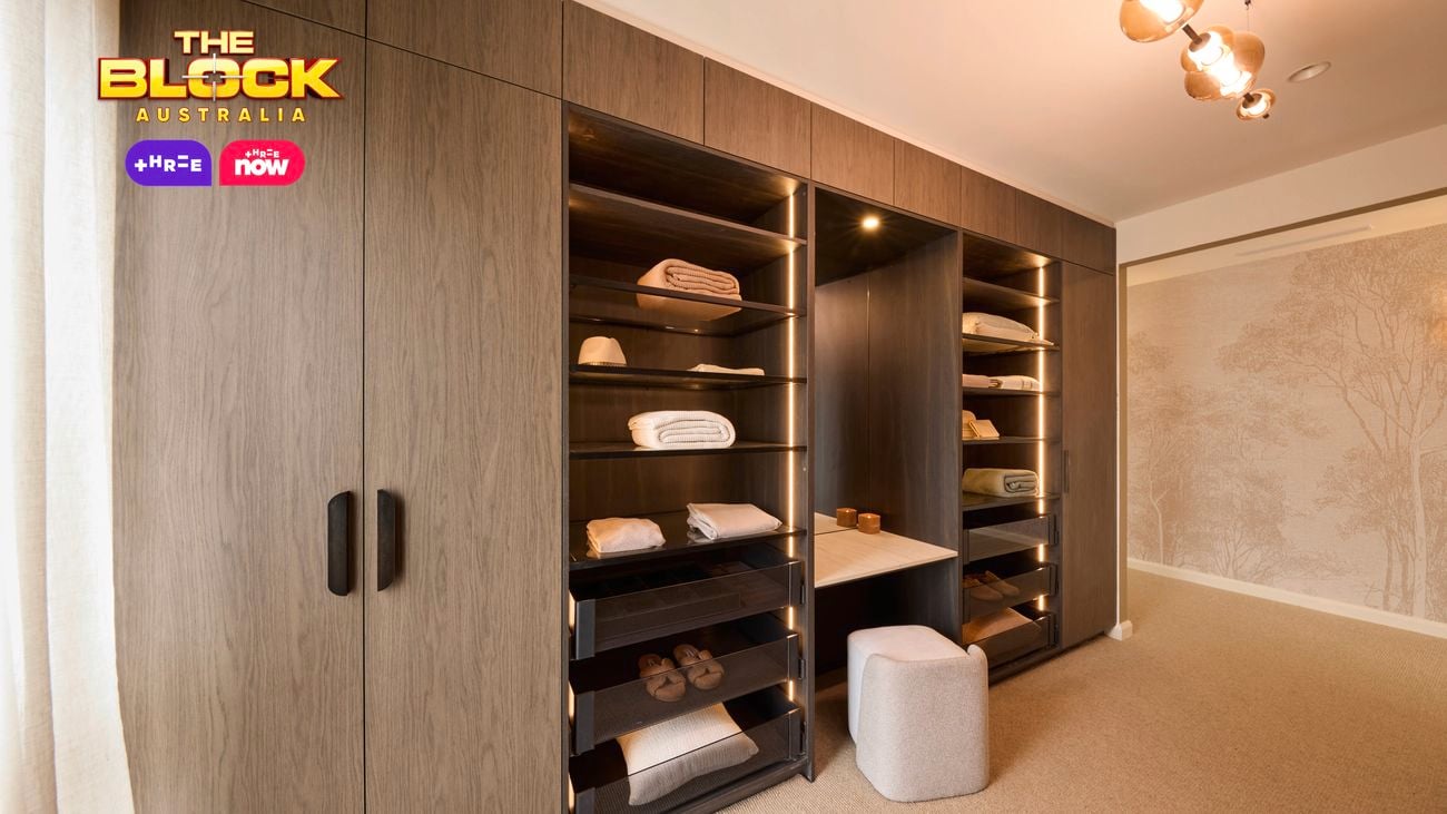 A well-organized walk-in closet featuring wooden cabinetry on both sides. The left side has closed cabinets with handles, while the right side displays open shelves with neatly folded clothes and towels. A small cushioned stool sits near the shelves on a carpeted floor. The space is illuminated by ceiling lights and natural light from a window at the far end. 