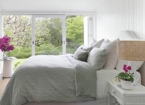 A bright bedroom with a bed a decorative full-length mirror on the wall and a large window looking out onto a lush green garden.