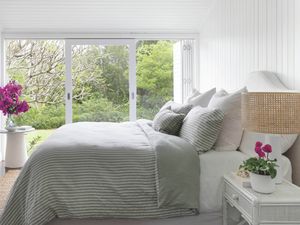 A bright bedroom with a bed a decorative full-length mirror on the wall and a large window looking out onto a lush green garden.