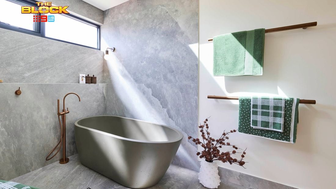 Modern bathroom with matte gray freestanding tub, stone-look gray tiles, wall-mounted faucet, green towels on wooden racks, and natural light from a high window. 
