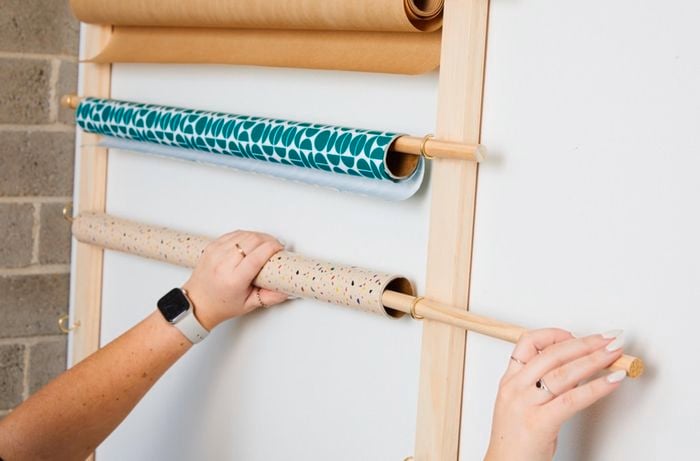  A pair of hands sliding wooden dowels onto hooks attached to a wall. Different coloured rolls of wallpaper are hanging on the dowels