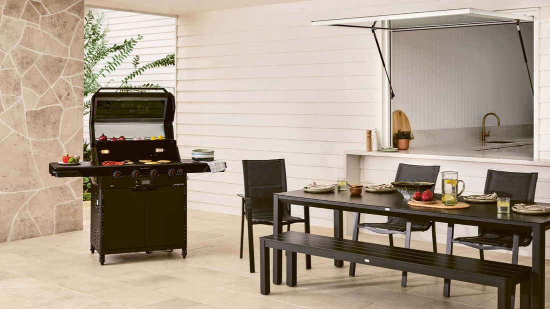  A black barbecue grill with its lid open, cooking onions, bell peppers and flatbreads on the griddle, set on a patio next to a dining table and chairs.
