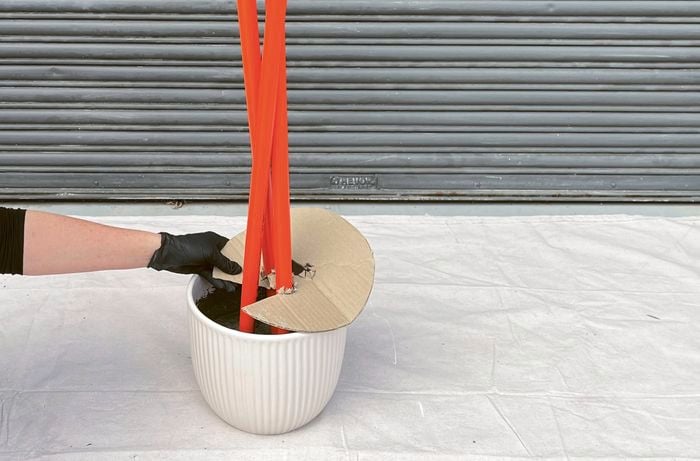 A gloved hand positioning a cardboard jig on a white pot holding orange conduits set into concrete, against a corrugated metal background.