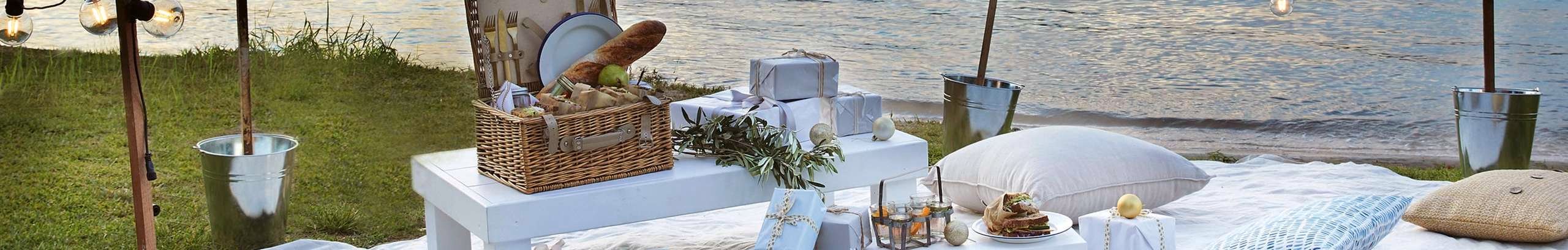 picnic setup with basket, blanket, cushions and christmas gifts on the grass near water