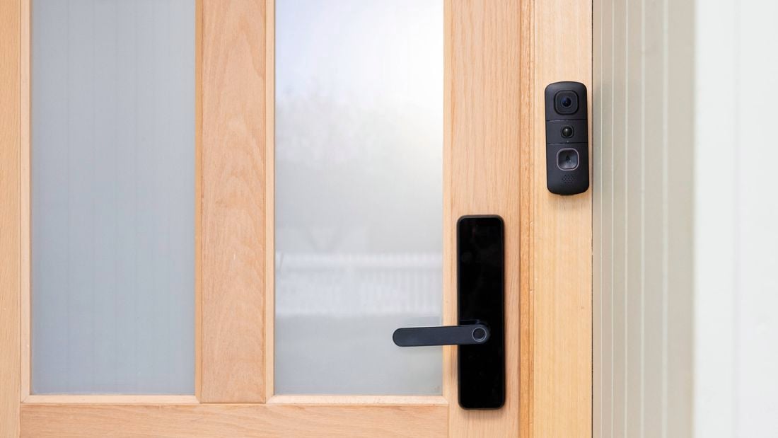 A black Swann video doorbell is mounted beside the handle of a wooden front door with frosted glass panels. 