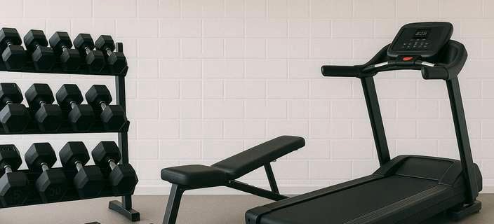 Home gym setup with a treadmill, weight bench, and dumbbell rack against a white brick wall