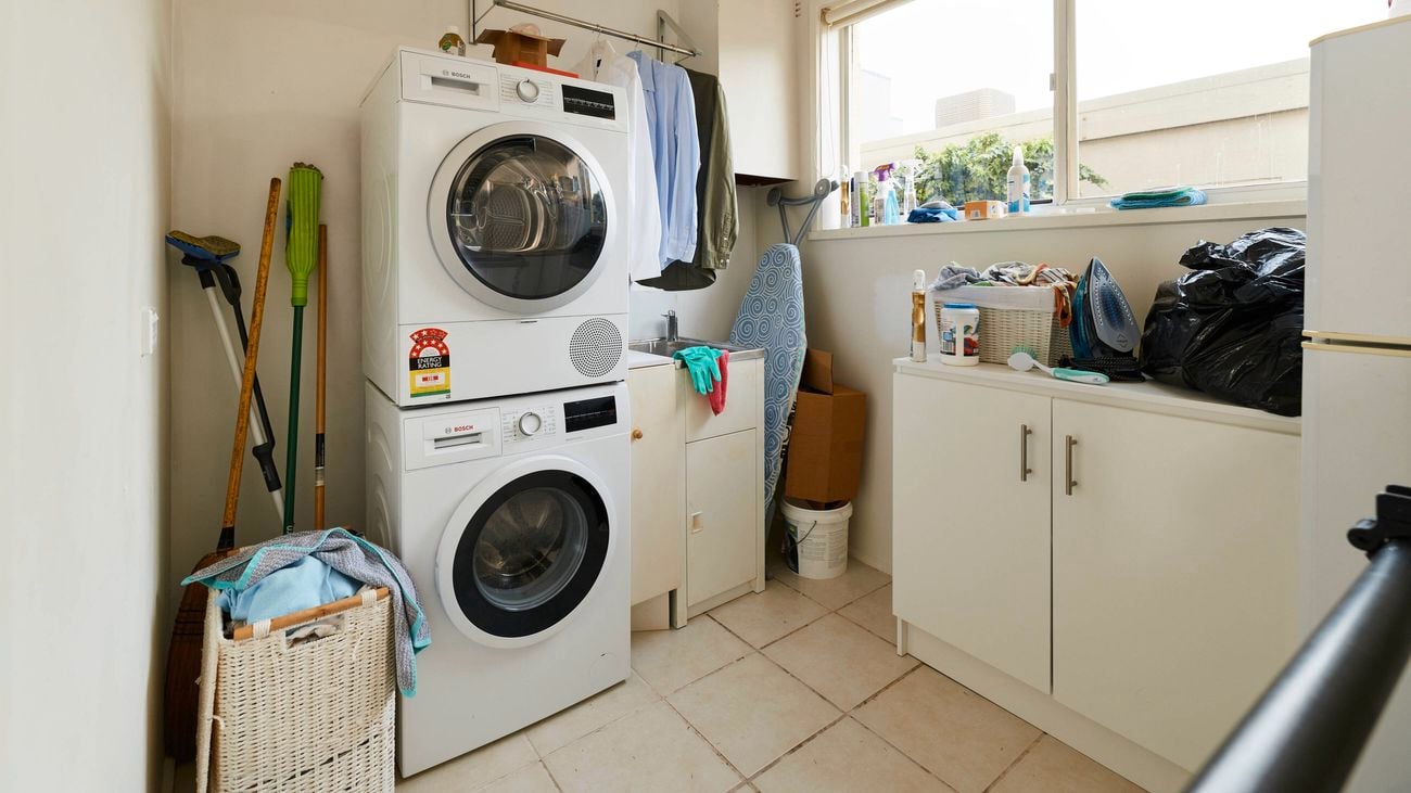 Compact laundry with stacked front-loading washer and dryer beside a sink, hanging clothes on a rail above, cleaning items on a benchtop, and a large window providing natural light.