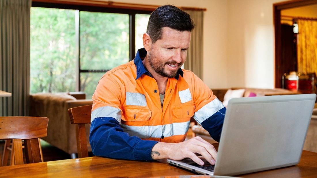 A man in hi-vis working on a laptop placed on a dining table inside a home