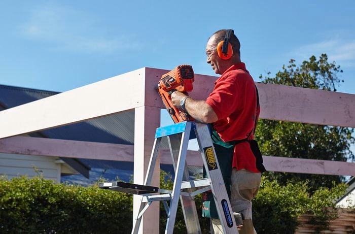 A person on a ladder attaching a beam to a post using a nail gun