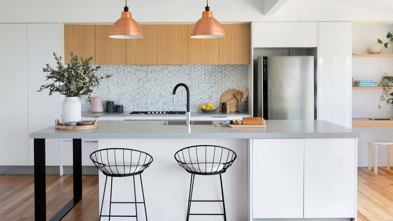 Modern kitchen with white cabinetry, light timber overhead cabinets, hexagonal tiled splashback, black chairs, copper pendants and black and stainless steel tapware.