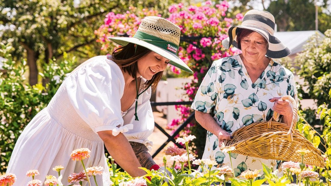 Katie of Little Lyndoch Flower Farm and her mother wear sunhats while gardening outdoors among pink and orange flowers.