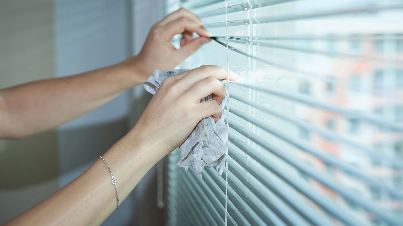 A pair of hands using a cloth to wipe between the slats of a set of vertical blinds